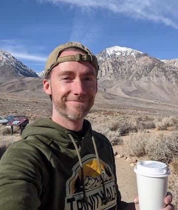 Photo of Greg Moyerbrailean smiling, holding a coffee cup in front of a mountain range on a sunny day.